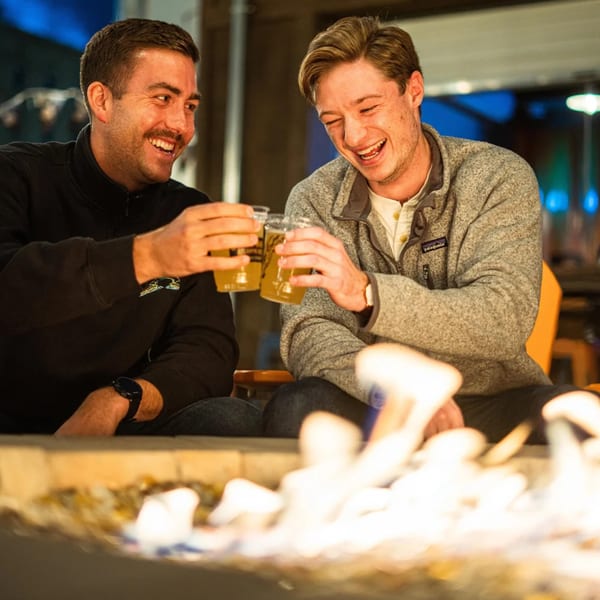 Two men laughing and toasting beers by a fire pit at Tennessee Avenue Beer Hall.