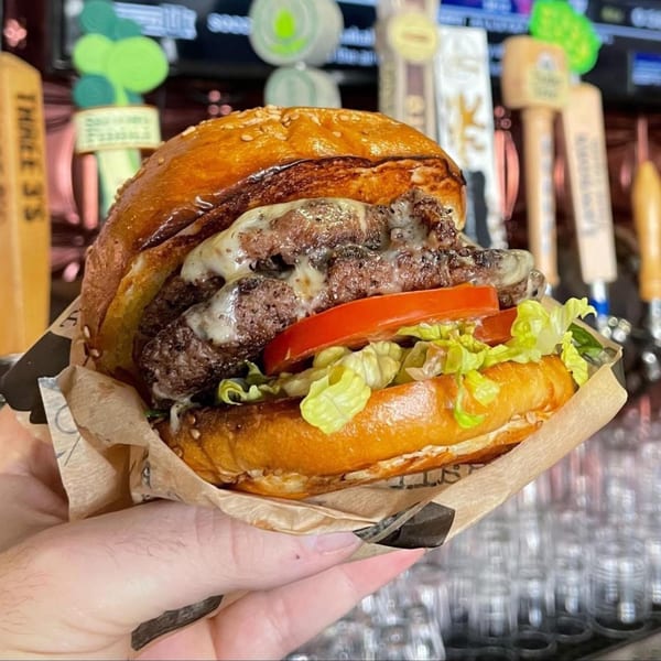 A hand holds a Tennessee Avenue Beer Hall burger with melted cheese, lettuce, and tomato.