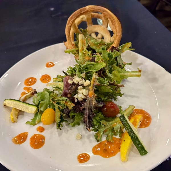 A fresh salad with mixed greens, cherry tomatoes, zucchini, and a pretzel crisp on a white plate.