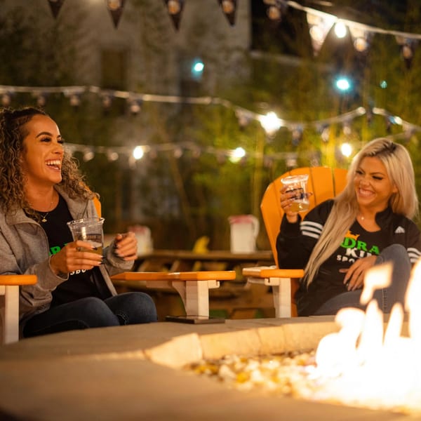 Two women laughing by a fire pit at night, holding drinks, with string lights overhead.