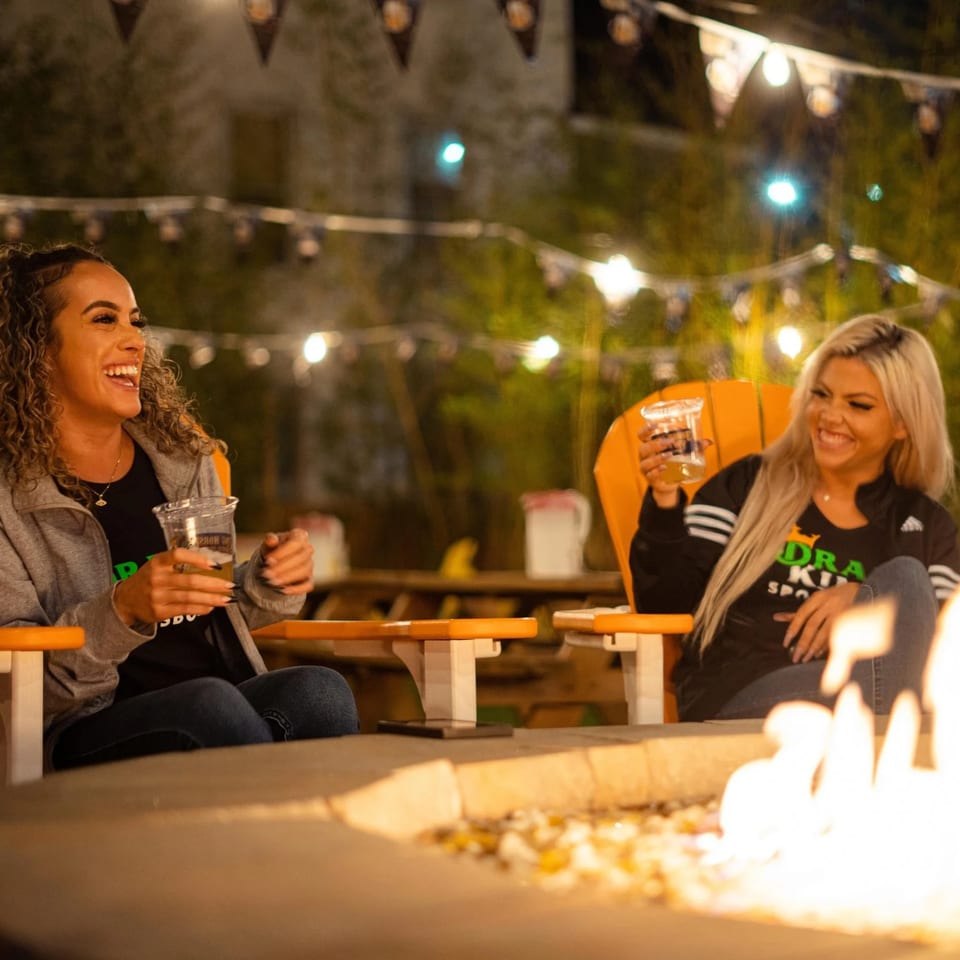 Two women laughing by a fire pit at night, holding drinks, with string lights overhead.