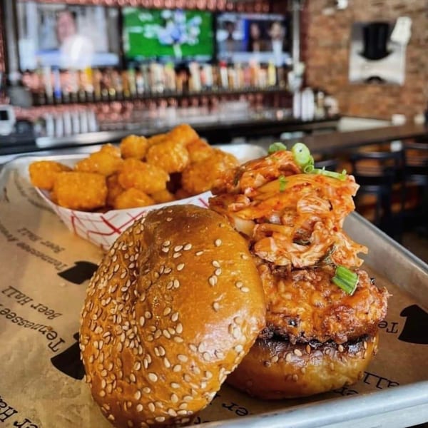 A Tennessee Avenue Beer Hall burger with tater tots on a metal tray, with a bar in the background.