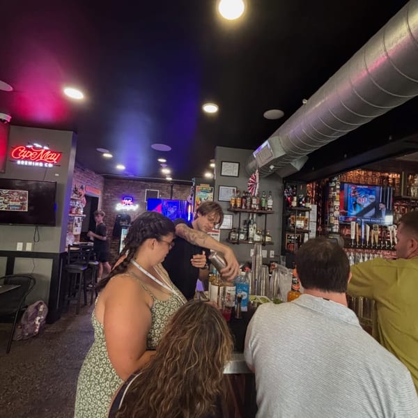 Bartender prepares drinks for customers at a busy bar with a Cape May Brewing Co neon sign.