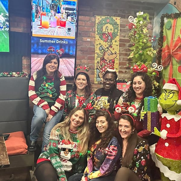 Seven smiling women in ugly Christmas sweaters pose in a festive bar with holiday decorations.