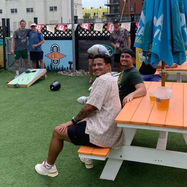 Two men sit at an orange picnic table with beer, while others play cornhole on artificial grass.