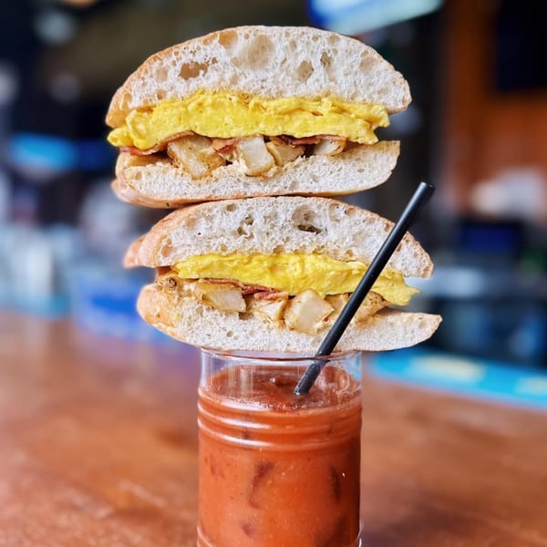 Breakfast sandwich with scrambled eggs, bacon, and potatoes, balanced atop a Bloody Mary cocktail.