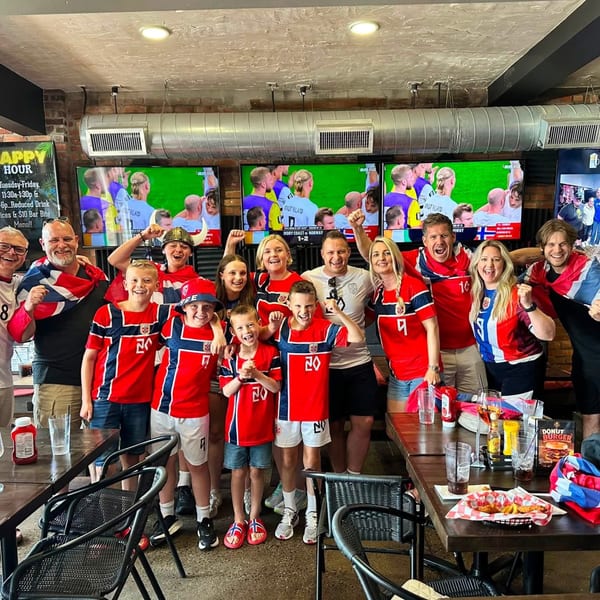 Cheerful group of adults and children in Norwegian gear watching a soccer game in a lively restaurant.
