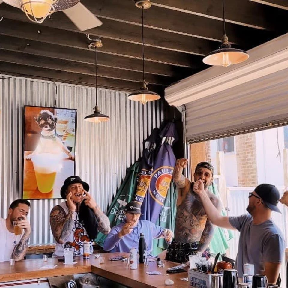 Five men laughing and drinking at a bar, with a large beer poster in the background.