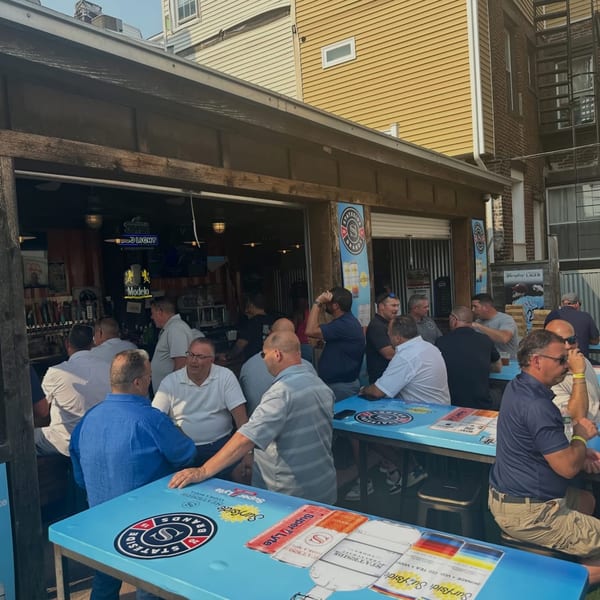 Patrons gather at an outdoor bar with branded blue tables and a Modelo sign.
