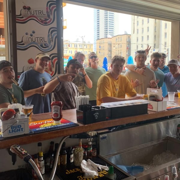 Young men pose behind a bar with drinks and cigars, featuring NÜTRL branding and city views.