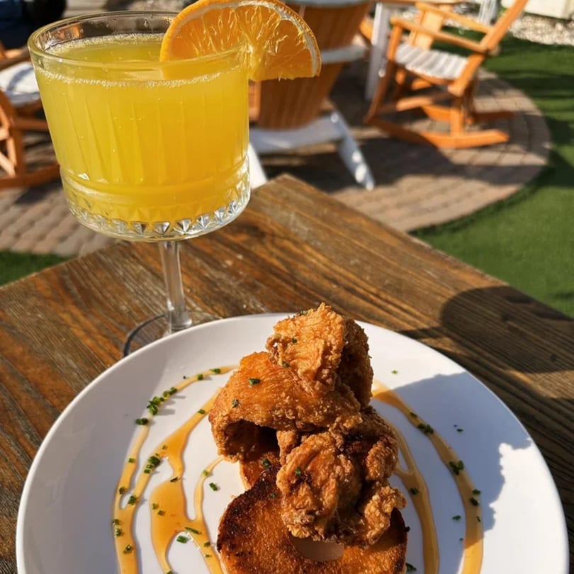 Fried chicken and pancakes drizzled with sauce, alongside an orange cocktail on a wooden table.
